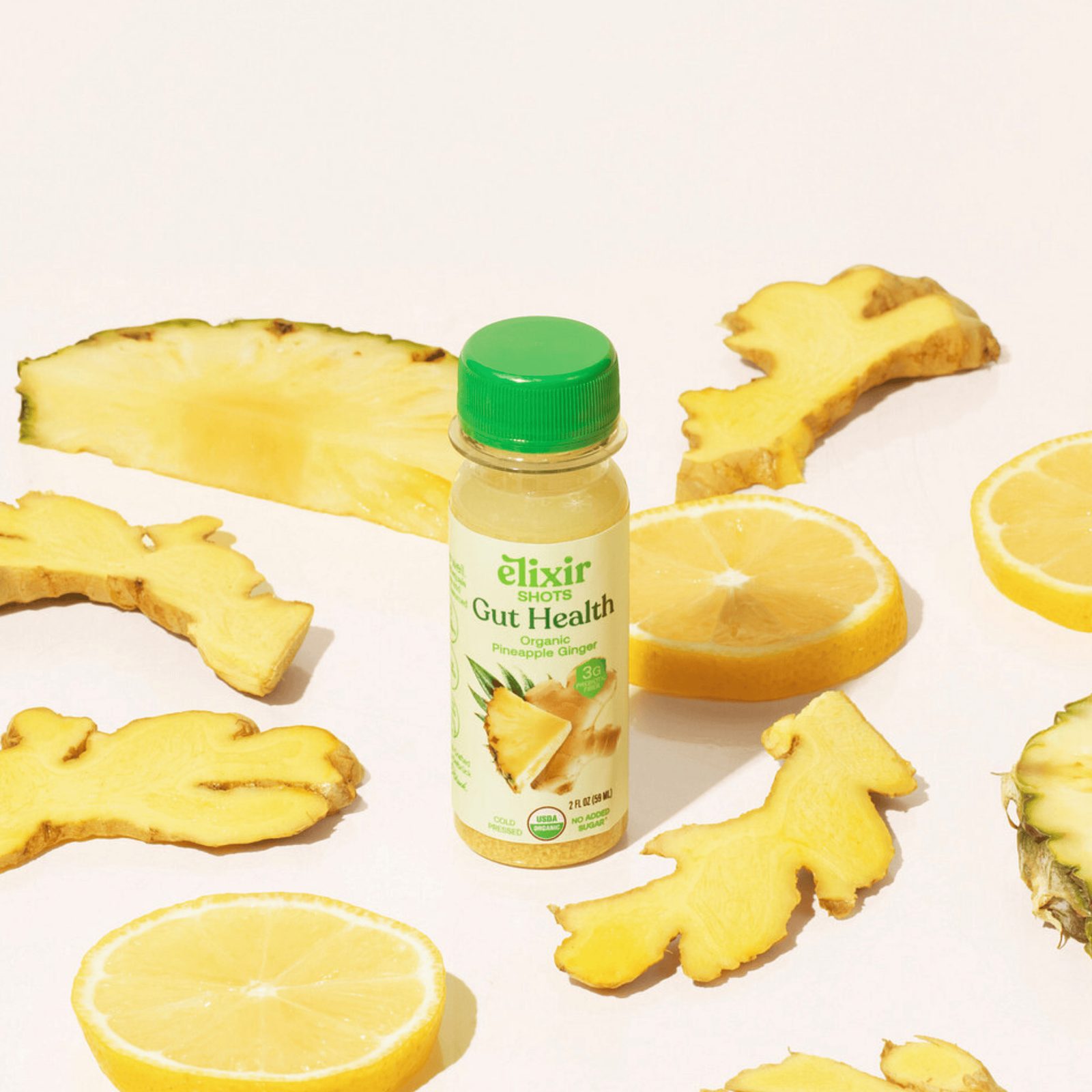 An Elixir Gut Health shot beside fresh ginger, lemon and pineapple